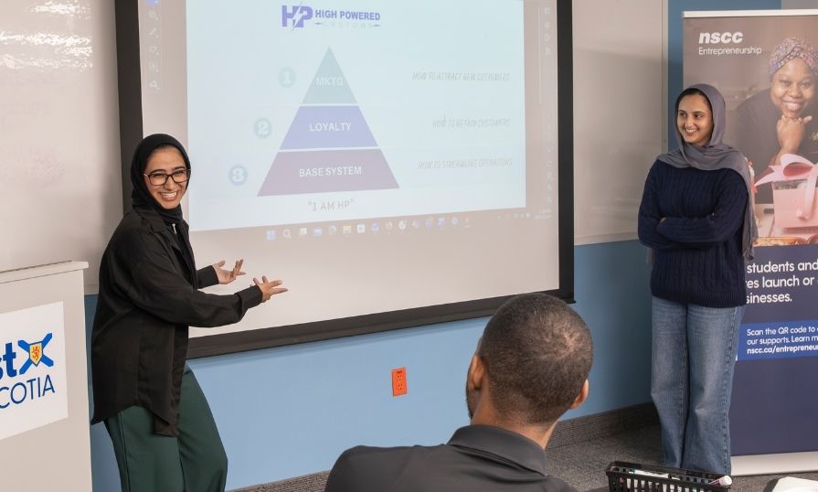 Two smiling students presenting to one person, with a presentation screen in the background.