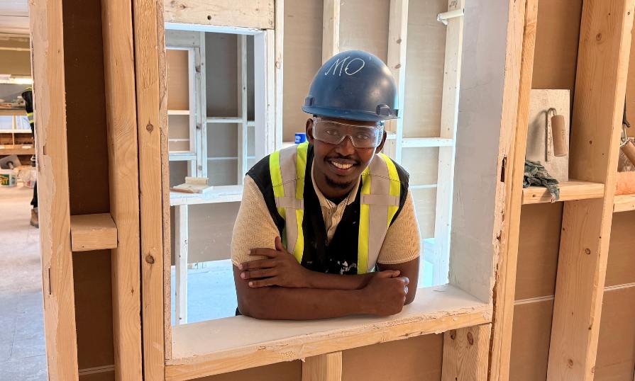 Man smiling in carpentry workshop in window frame wearing safety gear.