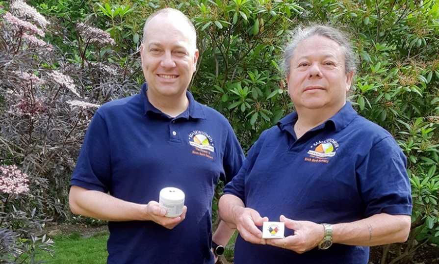 Two men stand in front of trees holding birch bark skincare products.