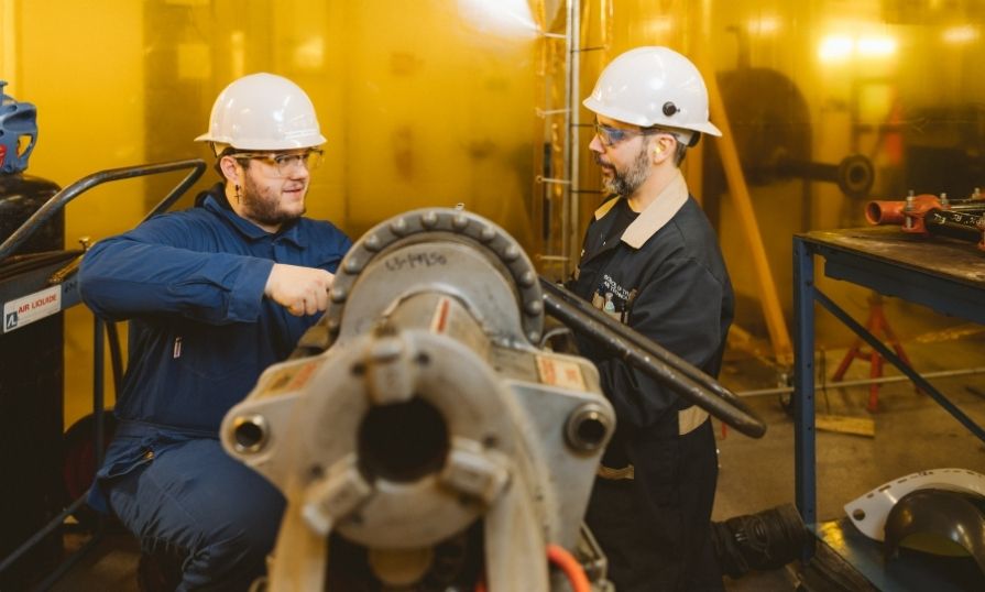 Two people wearing safety PPE and hard hats work on a large pipe in a workshop.