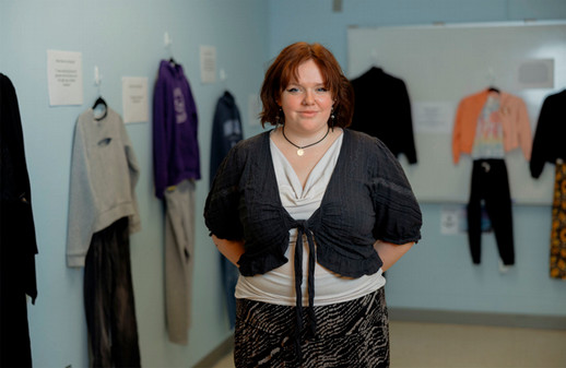 NSCC student Mia stands in the classroom, in front of her exhibit with clothing hung on the walls behind her.