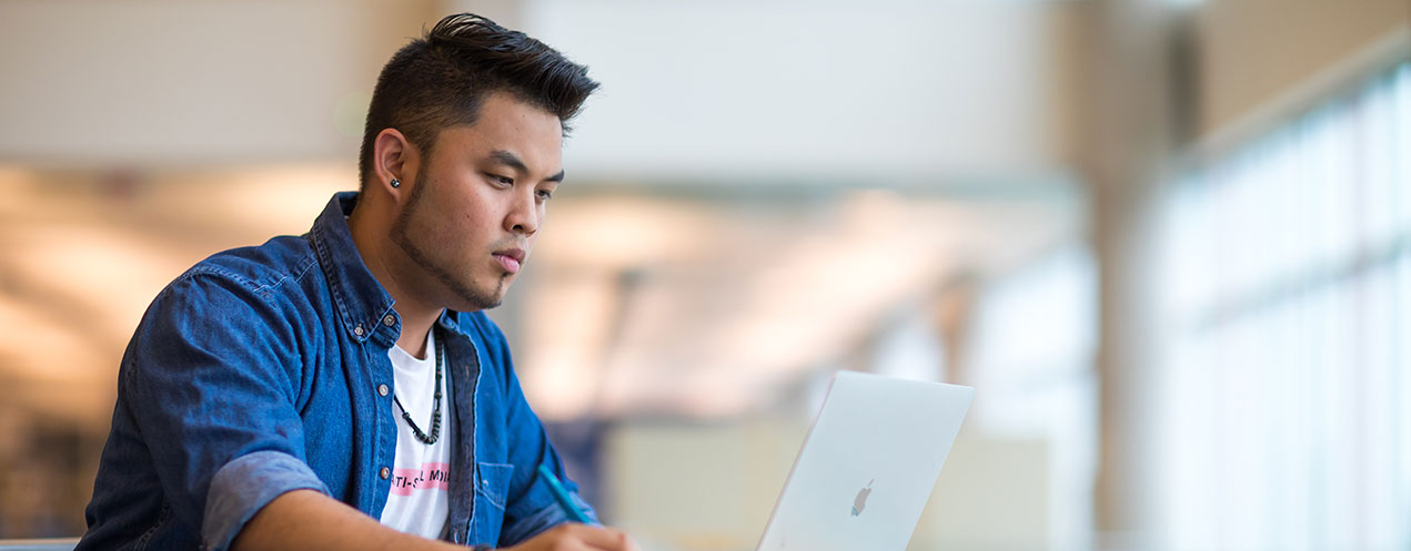 A person wearing an open, button-down shirt over a t-shirt sits in front of an open laptop and looks at the screen while holding a pen in their right hand.