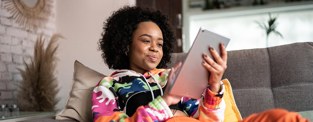 A person wearing a colourful hooded sweatshirt sits on a couch with a cushion behind their back while holding and looking at a tablet. 