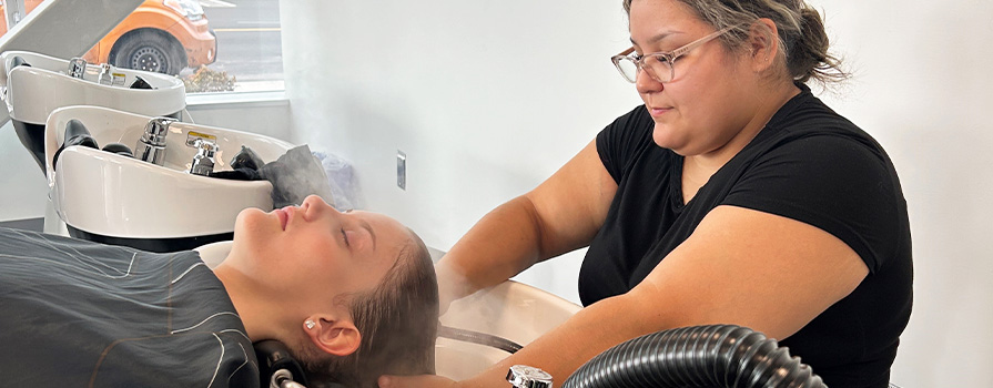 A student gently rinses the hair of another student while their head rests at the sink of a salon.