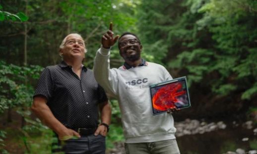 Two people in the woods. One is pointing up towards something while holding an iPad showing wildfire simulation data.