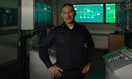 Jay Julien wears a uniform and stands indoors on a ship with his hands on his hips.