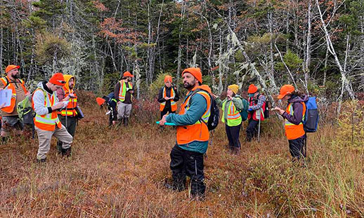 A group of NRET students wearing safety vests and toques learn in a wooded area.