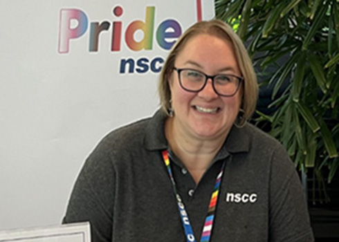 A portrait of NSCC 2SLGBTQ+ Student Services advisor, Melanie Karas. She is seated at an information booth table and is in front of a sign that reads Pride NSCC.