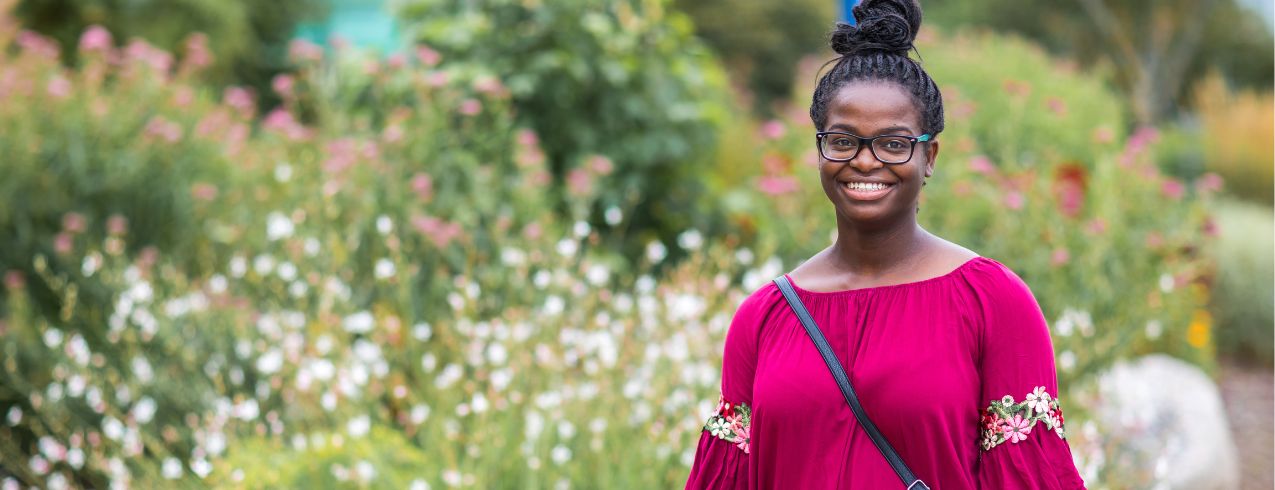 A Black woman smiling at the camera in front of a flowering bush.