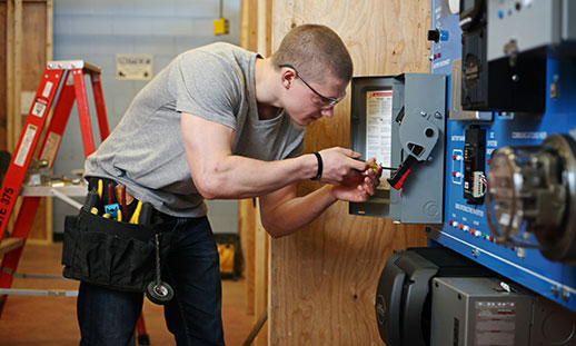 A student wearing a toolbelt uses tools to work on an electrical panel.