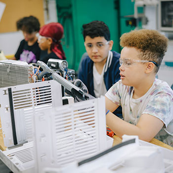 Four youth sit together at a table in a workshop setting and learn about equipment.