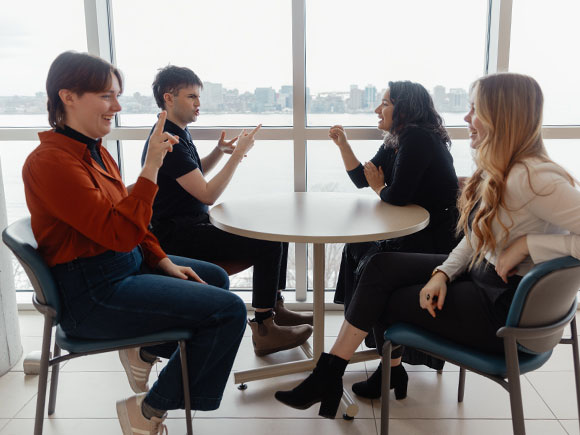 Four ASL students laugh while sitting in front of a window overlooking the Halifax Harbour signing to one another.