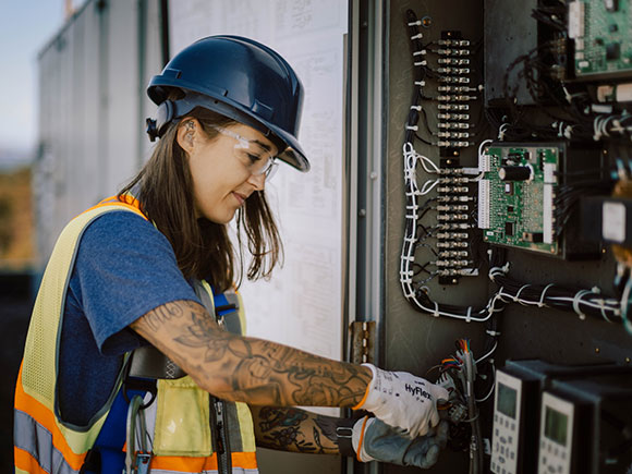 An HVAC professional wearing a safety hat, vest, glasses and gloves looks at wiring.