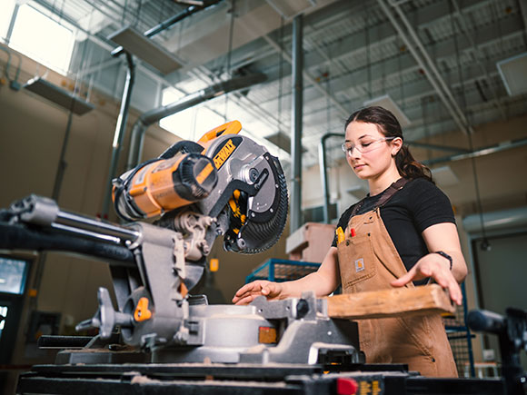 A student stands in front of a power saw and prepares to cut a piece of wood.