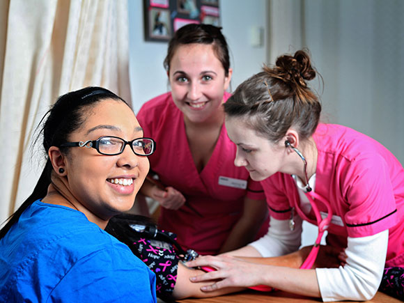 Three students in brightly coloured scrubs practice taking blood pressure readings.