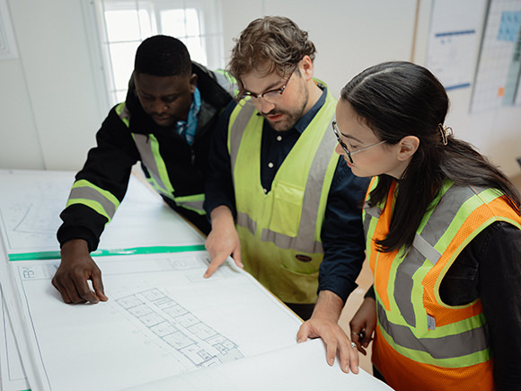 Three students in safety vests look at blue prints on a table.