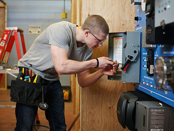 A student wearing a toolbelt and safety glasses uses a screwdriver in a wall-mounted unit.