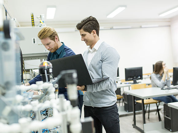 Two men work in a lab or classroom environment; one of the men is holding a laptop computer.