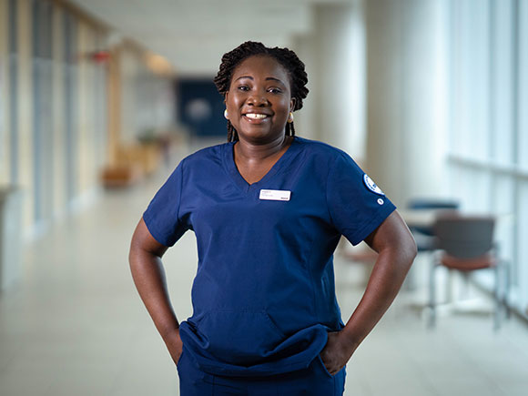 A nursing student stands facing the camera and smiling. She is wearing blue scrubs.