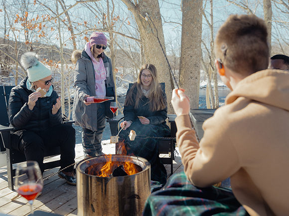 A group of students are seen gathered around a campfire in a wintery woods setting, roasting food and tasting wine.