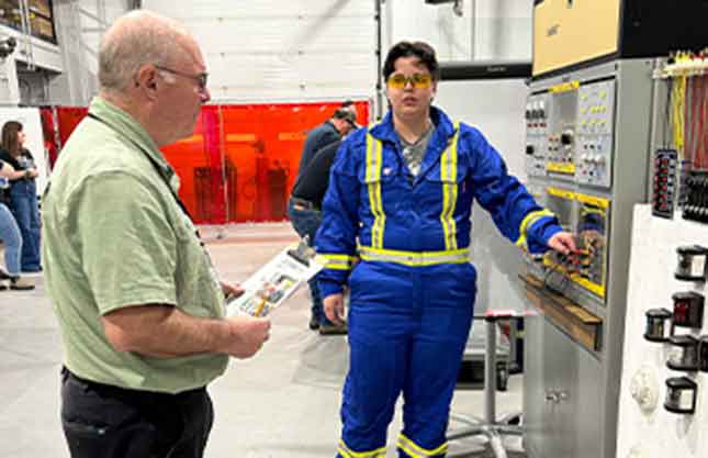 A student in protective glasses and reflective, blue coveralls works on a machine in a lab setting with faculty overseeing the work.