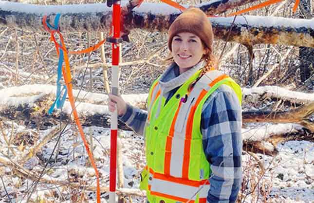 A smiling woman in a reflective vest and brown toque works in the snowy woods.