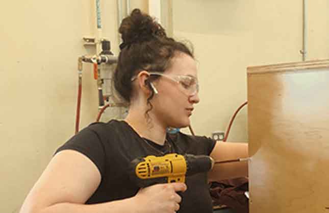 Profile image of a woman in safety glasses and ear plugs drilling into a piece of wood in a workshop setting.