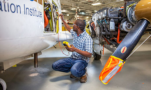 An aircraft maintenance diploma student works beside the body of an airplane holding a piece of machinery.