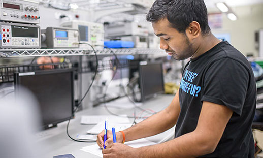 A student stands at a long desk covered in monitors, wires and other electronic engineering equipment.