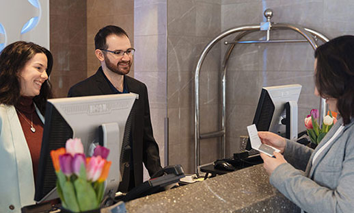 Two Hotel and Restaurant Management students stand behind a hotel reception desk and speak with a guest.