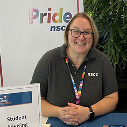 A portrait of NSCC 2SLGBTQ+ Student Services advisor, Melanie Karas. She is seated at an information booth table and is in front of a sign that reads Pride NSCC.