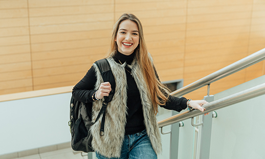 A college student wearing jeans, a turtleneck sweater and vest climbs a staircase and smiles at the camera. She carries a backpack over her right shoulder.