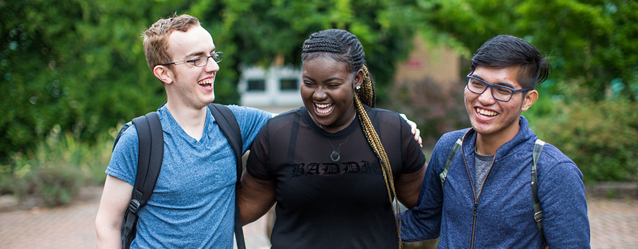 Three smiling college students walk together outdoors in an NSCC campus courtyard with their arms around one another’s backs.
