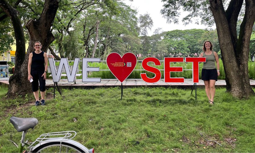 Two women stand by a “WE Love SET” sign in a green park with trees