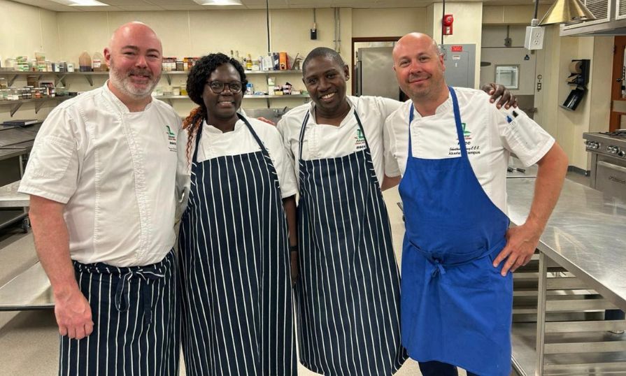 Lynette Nyagaya with chefs in a professional kitchen, smiling.