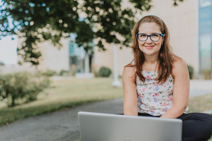 Student sitting in front of campus with open laptop.