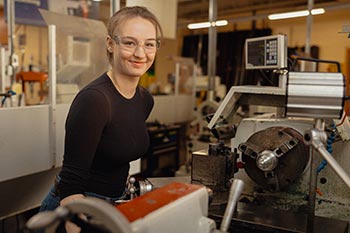 NSCC Precision Machining graduate Mollie smiles toward the camera in the shop classroom. She is wearing a dark long sleeved shirt and protective safety glasses. 